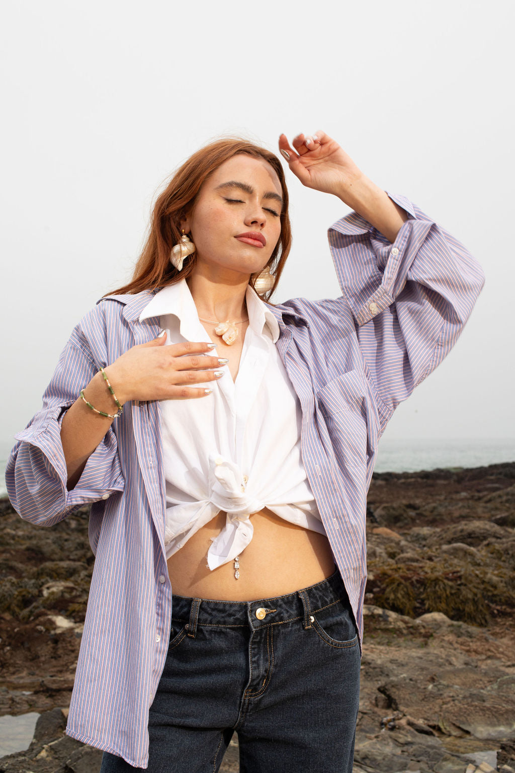 Model with eyes closed, resting one hand on her chest and delicately raising the other over her eyes, wearing the Agnes turbo shell earrings on 14K gold-filled ball posts, the Angaria Delphinus shell pendant silver necklace (Thalassa Necklace), and the Santiago wrap serpentine and pearl bracelet, with a coastal beach backdrop.