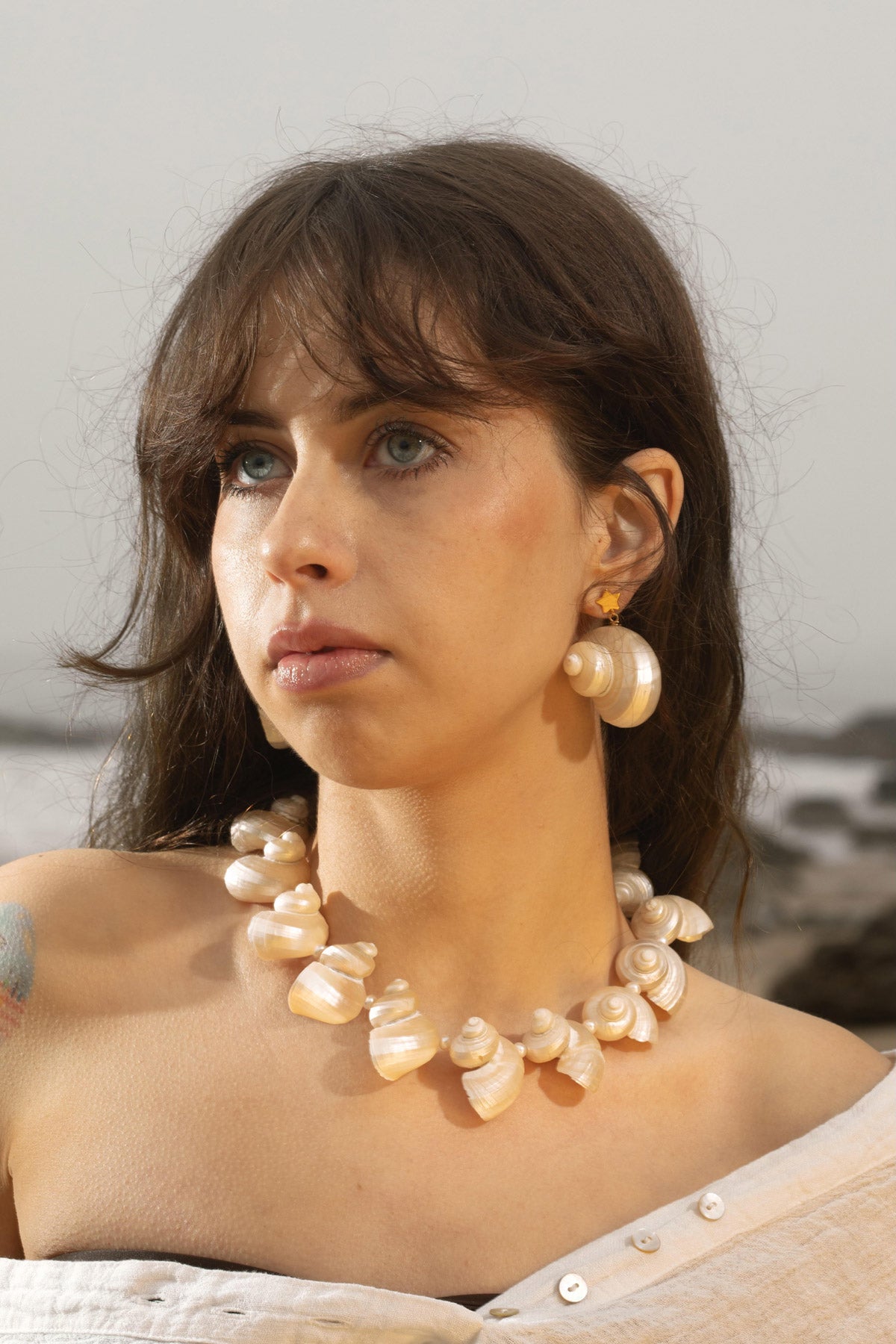 A model poses on a moody beach, her hair flowing in the wind. She wears the Astr Earrings, gold-plated star studs with turbo seashell drops, paired with the Mer Necklace. The sculptural seashell and freshwater pearl necklace creates a modern, soft look, blending natural textures, organic shapes, and gentle imperfections into a cohesive design.