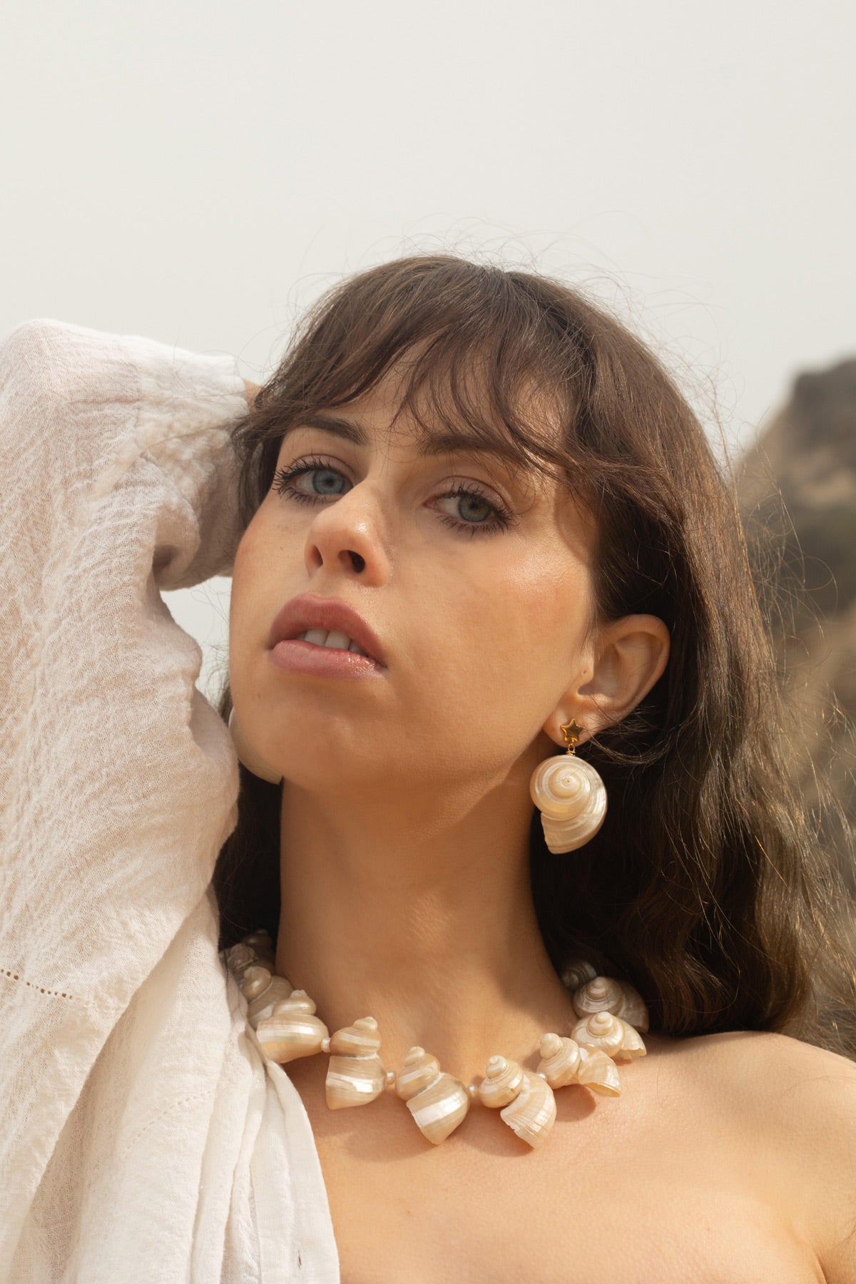 The model poses in front of a rocky hilltop, wearing the Astr Earrings and Mer Necklace. Her wind-blown hair frames the celestial dangle earrings, complementing the chunky turbo seashell necklace. Together, the pieces create a modern, feminine aesthetic that blends natural beauty with contemporary style.
