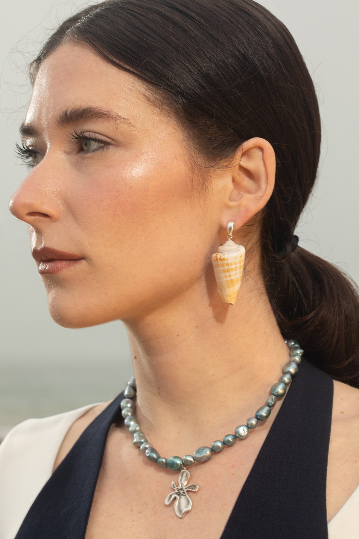 Model wearing the Bea Earrings with cone-shaped seashell sculptural drops, hair pulled back to highlight the earrings against the coastal sky. The Cleo Necklace, featuring a chunky blue baroque pearl and sterling silver flower pendant, is also visible.