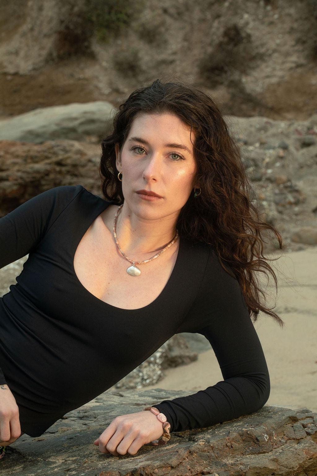 Model resting on a beach rock during golden hour, wearing the Leone Bracelet, a chunky ocean jasper beaded bracelet, and the Caroline Necklace, a jasper and freshwater pearl necklace with a sterling silver-plated clam shell pendant.
