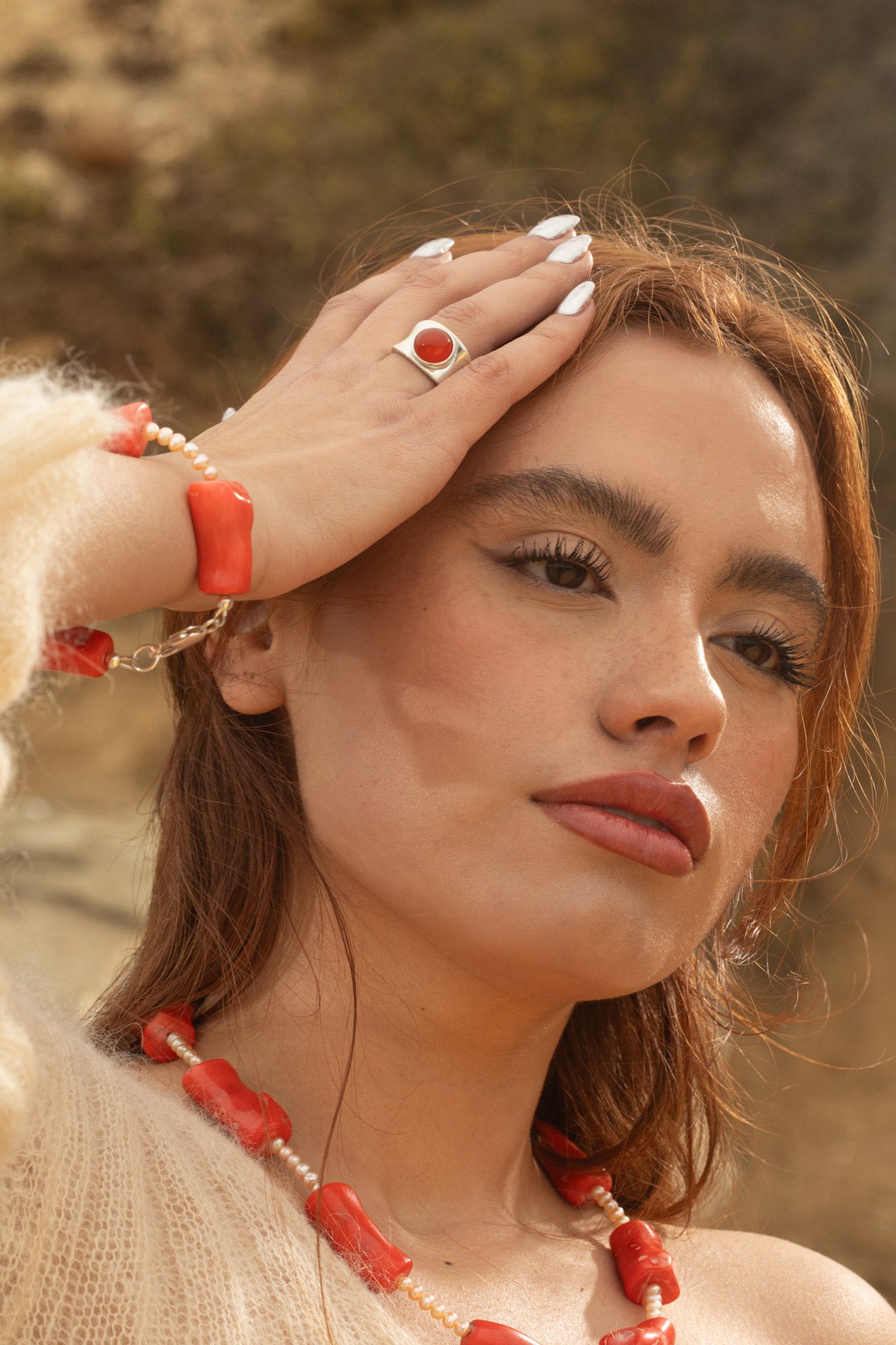 Model wearing the Coral Mini Bracelet with small freshwater baroque pearls and chunky coral beads, resting her hand on her head to show the bracelet. She’s also wearing the Alexandra Ring, a chunky sterling silver concave ring with a carnelian cabochon, and the Coral Necklace featuring coral and freshwater baroque pearls. The photo, taken during golden hour, captures a rocky coastline backdrop.
