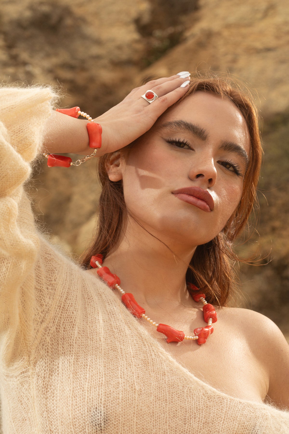 Model wearing the Coral Mini Bracelet with small freshwater baroque pearls and chunky coral beads, resting her hand on her head to show the bracelet. She’s also wearing the Alexandra Ring, a chunky sterling silver concave ring with a carnelian cabochon, and the Coral Necklace featuring coral and freshwater baroque pearls. The photo, taken during golden hour, captures a rocky coastline backdrop.