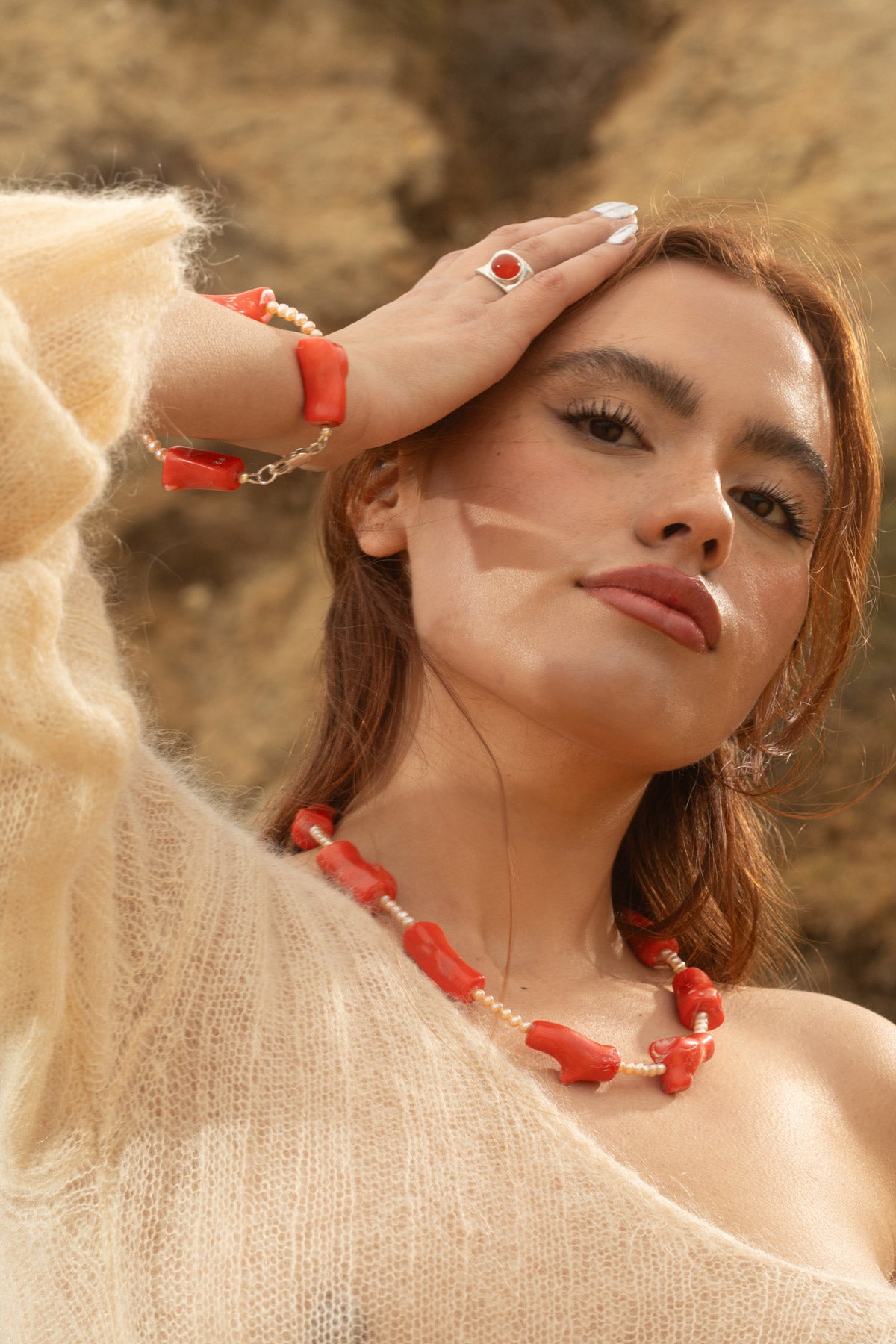 Model wearing the Coral Necklace and Coral Mini Bracelet, both featuring chunky coral beads and freshwater pearls, paired with the Alexandra Ring, a sterling silver concave ring with a carnelian cabochon. The rocky terrain backdrop highlights the natural textures and bold statement of the jewelry.