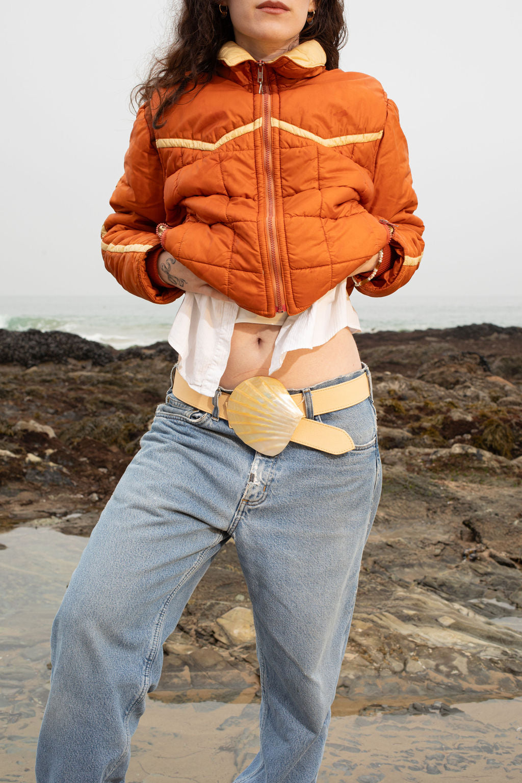 Model standing on the beach wearing jeans and an orange puffer, with her curly hair framing her face. The adjustable Emily Belt, with its mother of pearl buckle, stands out as a statement piece, showcasing its artisanal craftsmanship and modern, nature-inspired design. The ocean’s waves crash in the background, highlighting the belt’s organic beauty.