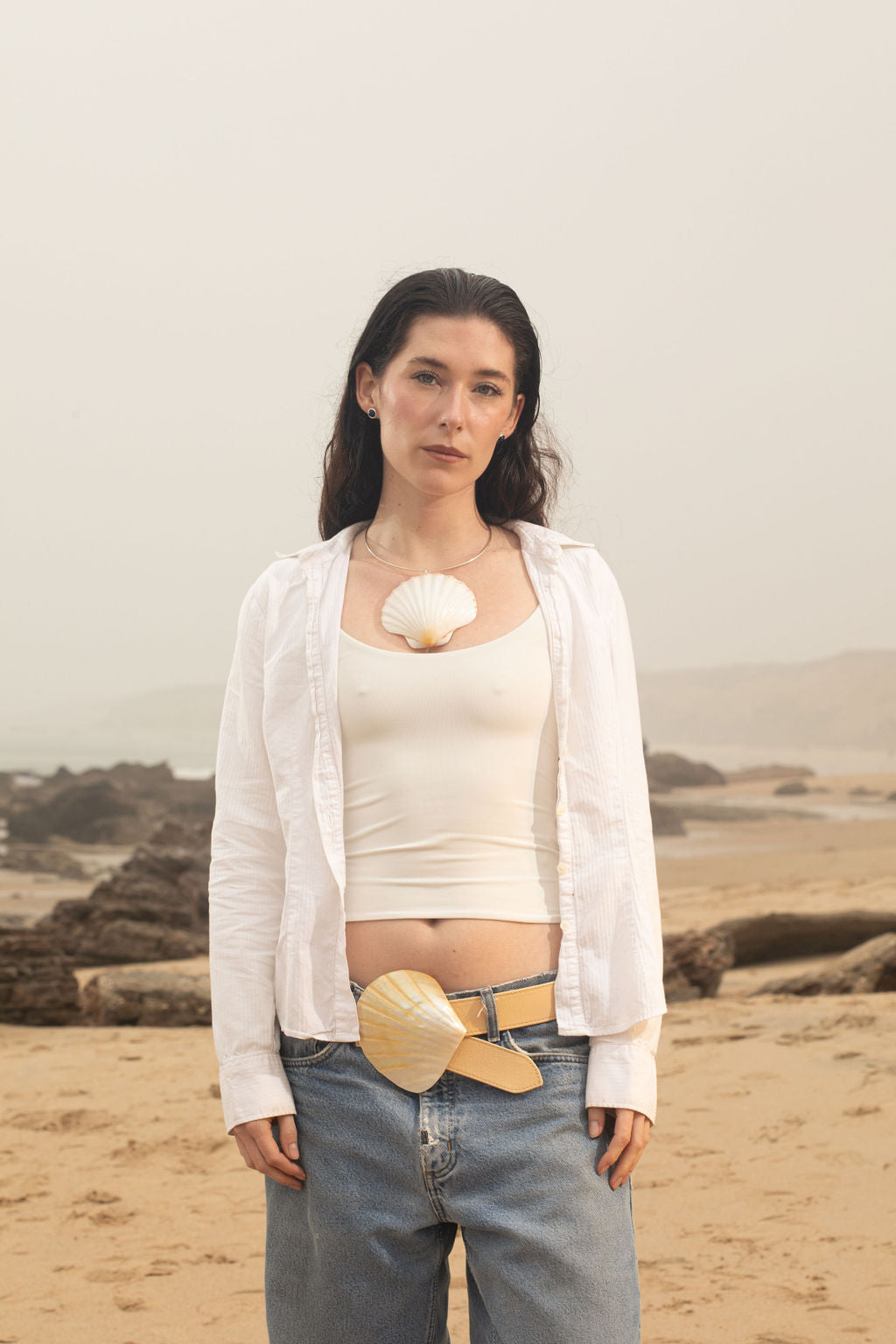 Model standing on the beach, wearing the Emily Belt with a mother of pearl buckle. The adjustable leather belt features expert craftsmanship and a nature-inspired, heirloom-quality design. Paired with the Marina Necklace, a sterling silver neck wire with a sculptural scallop shell pendant, creating a cohesive, organic, and modern look.