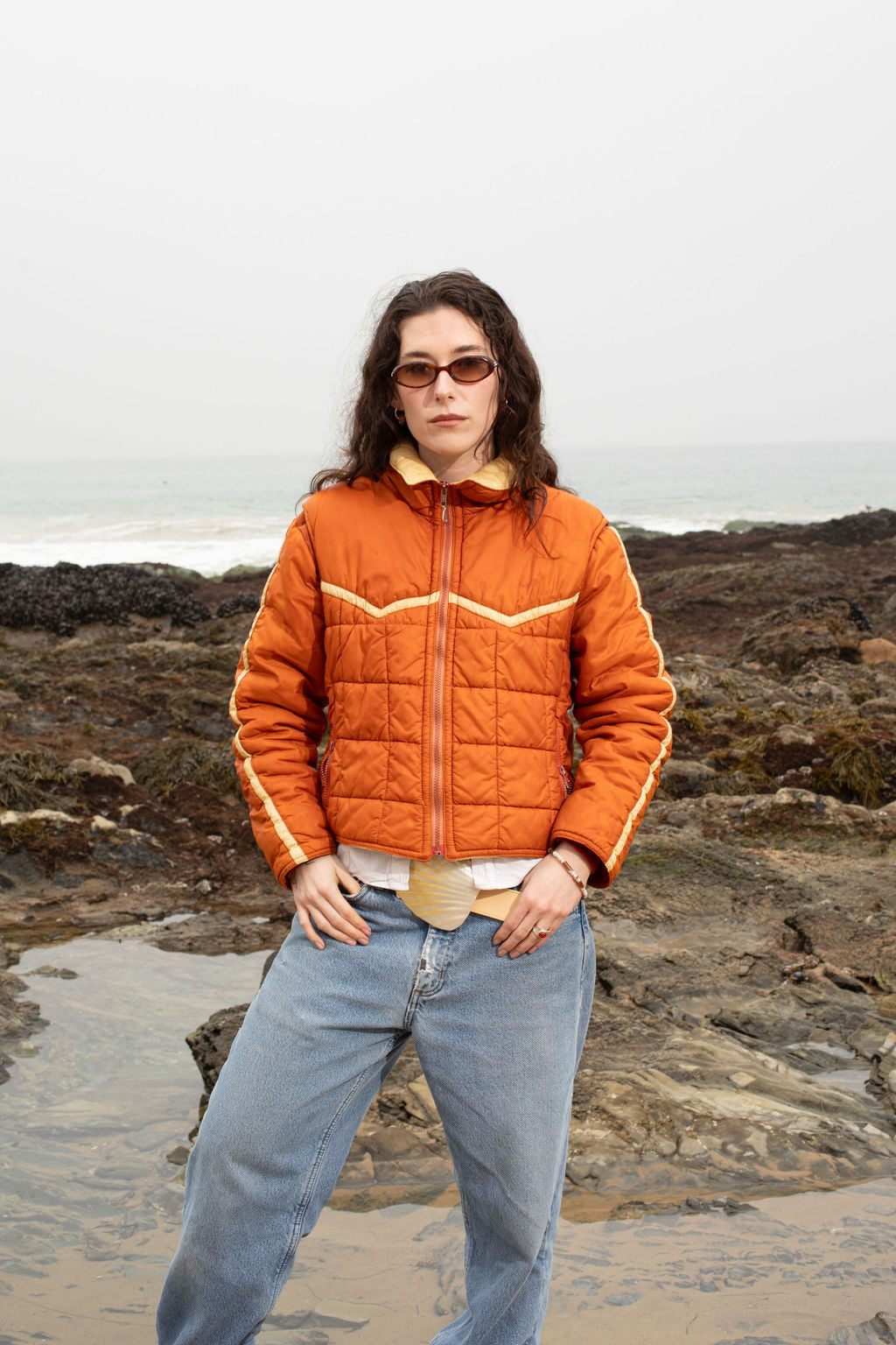Model wearing the Emily Belt with its adjustable cowhide leather and mother of pearl buckle, tucked under her orange puffer jacket. Paired with the Bennie Bracelet, a mix of freshwater pearls, agate, and jasper beads, and the Alexandra Ring, a sterling silver concave piece with a carnelian stone. Nature-inspired and handmade in Los Angeles.