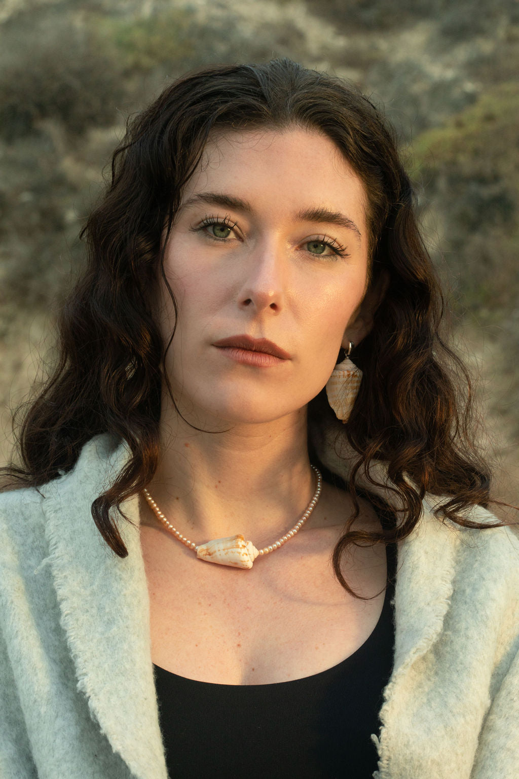 Model sitting on a rocky beach, staring into the camera with wavy, wet hair. The Isla Necklace features freshwater baroque pearls and a conch shell pendant, while the Bea Earrings showcase matching conch shell drops, creating a perfect coastal-inspired jewelry look.