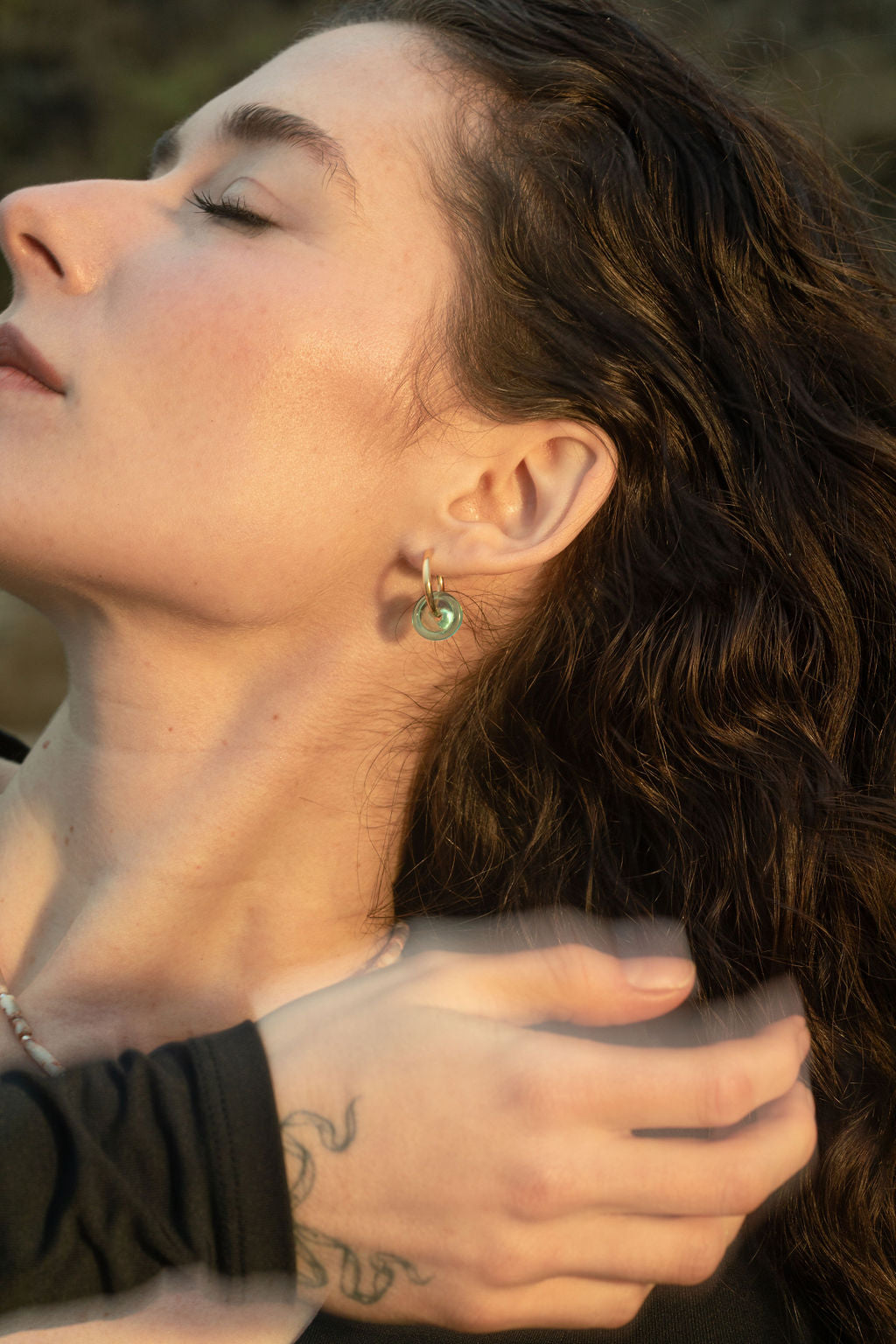 Close-up of model wearing the Keeley I Earrings as her hand, with a delicate bow tattoo, brushes past her ear. The turquoise glass beads glow against her skin, highlighting the minimal gold hoops.