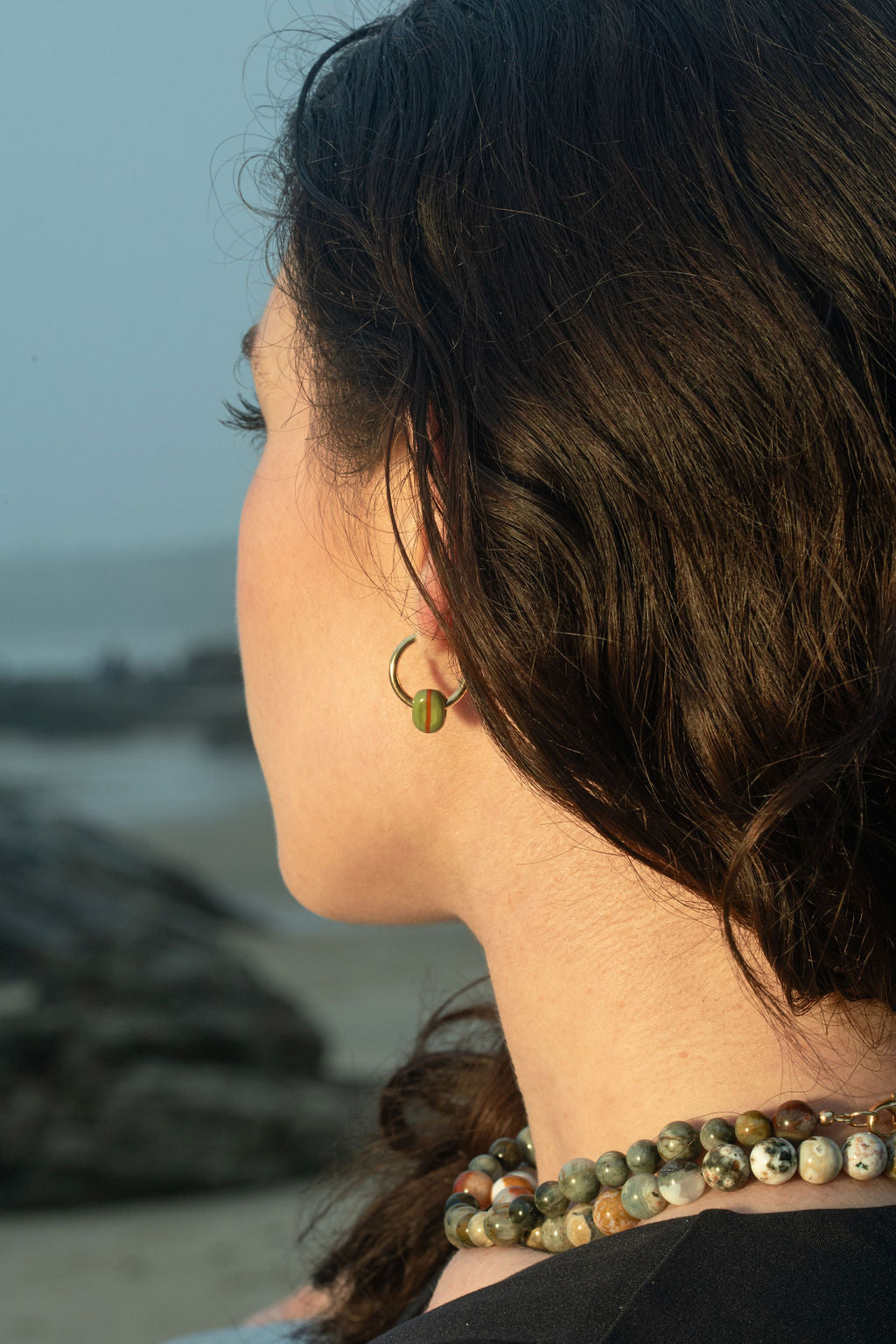 A close-up of the model wearing the Keeley II Earrings, olive handblown glass beads on gold-plated hoops, as she gazes at the beach during sunset. She’s also wearing the Pascal Ocean Jasper and Santal Eagle Eye Quartz beaded necklaces, layered together, creating an effortlessly chic, nature-inspired look.