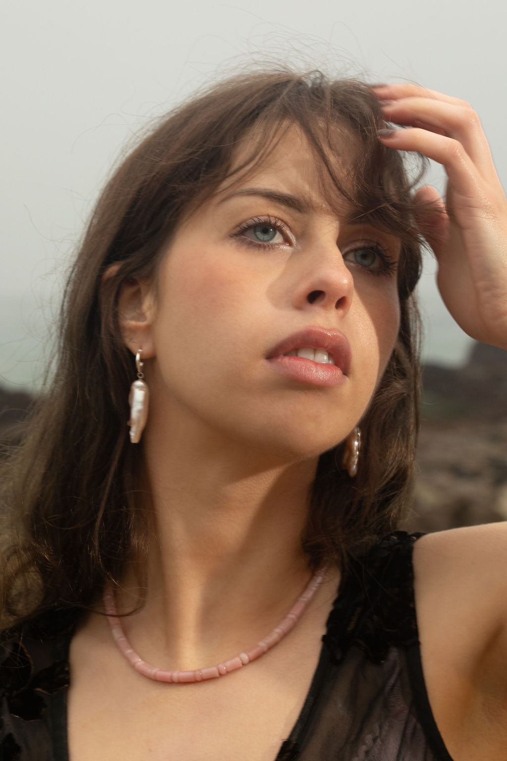 Close-up of a model wearing the Lottie Necklace and Peggy Earrings. Her hand gently touches her face, showcasing the organic beauty of the jewelry. Soft lighting and a natural coastal backdrop emphasize the warm tones of the pink opal necklace and freshwater pearl earrings.