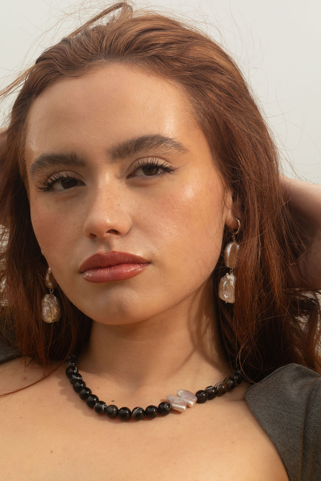 Model looking towards the camera, wearing the Sable Necklace with Black Sardonyx Agate beads and a baroque pearl pendant, and Siren Earrings in sterling silver 3/4 hoops with keshi and white rectangle baroque pearls. She’s pulling her hair back with her hands, showcasing both pieces of artisanal jewelry.