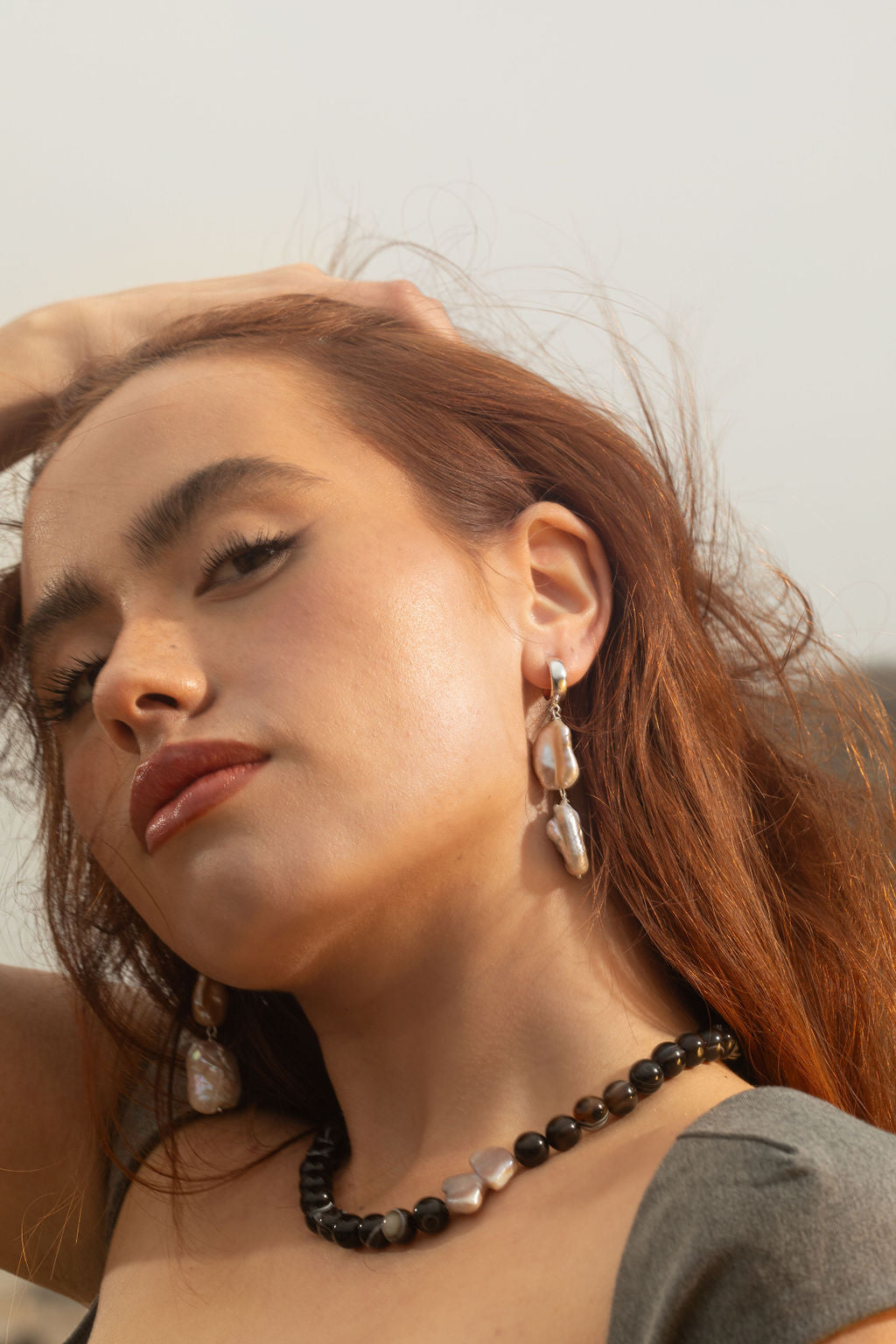 Model wearing the Sable Necklace with Black Sardonyx Agate beads and a baroque pearl pendant, viewed at an angle to highlight the side of the necklace. She’s resting her hand on her head, hair windswept, looking into the camera. Also wearing Siren Earrings, dangle-style, in sterling silver 3/4 hoops with keshi and white rectangle baroque pearls.