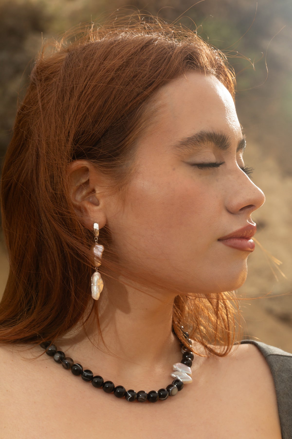 Side view of model wearing the Sable Necklace with Black Sardonyx Agate and a baroque pearl pendant, and Siren Earrings in sterling silver 3/4 hoops with keshi and white rectangle baroque pearls. Shot against a rocky coastal backdrop.