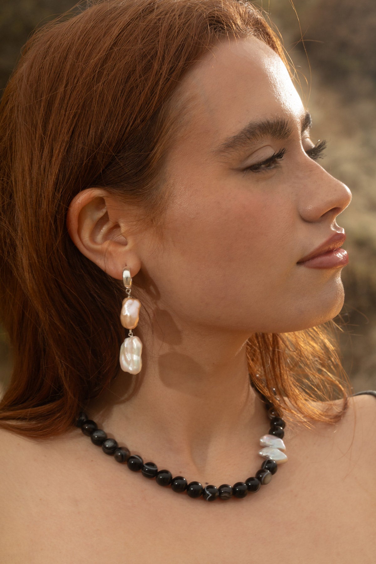 Model looking to the side, showcasing the Siren Earrings with tiered freshwater baroque and keshi pearls and the Sable Necklace featuring black sardonyx agate and an irregular baroque pearl pendant. The rocky coastline backdrop enhances the ocean-inspired look.