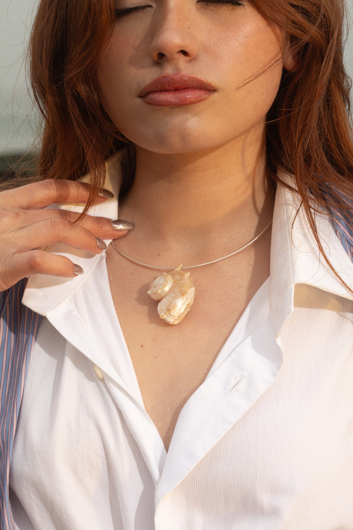 Close-up of model wearing the Thalassa Necklace, a handmade sterling silver cable chain with an Angaria delphinus seashell pendant. Windblown hair partially covers her face as she poses against a moody coastal backdrop, capturing the essence of ocean-inspired jewelry.