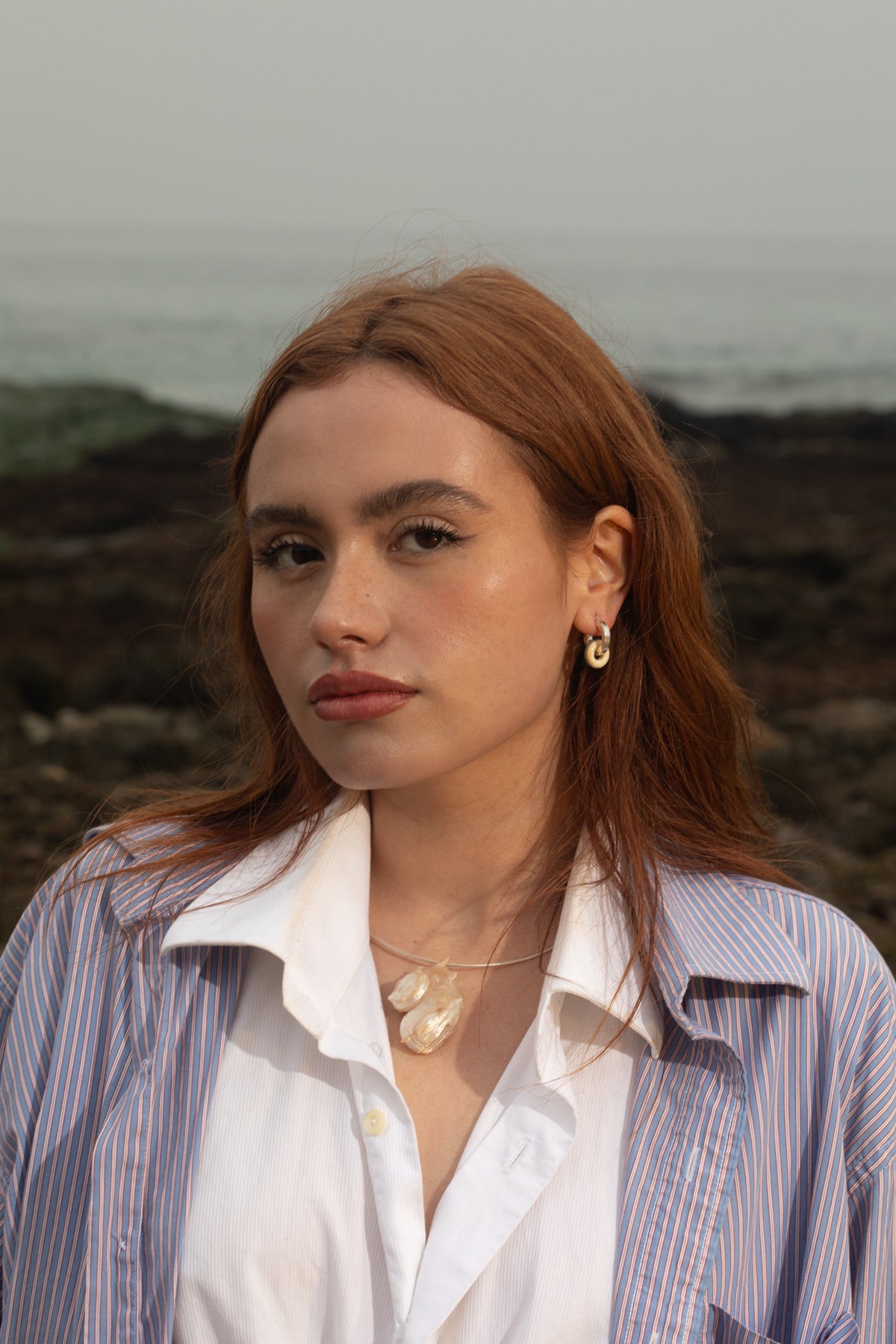 Model wearing the Thalassa Necklace, a piece of handmade jewelry featuring a sterling silver cable chain and Angaria delphinus seashell pendant, alongside the River Earrings—chunky sterling silver hoops with handblown glass beads. She looks into the camera at an angle, with a rocky, moody beach backdrop behind her.