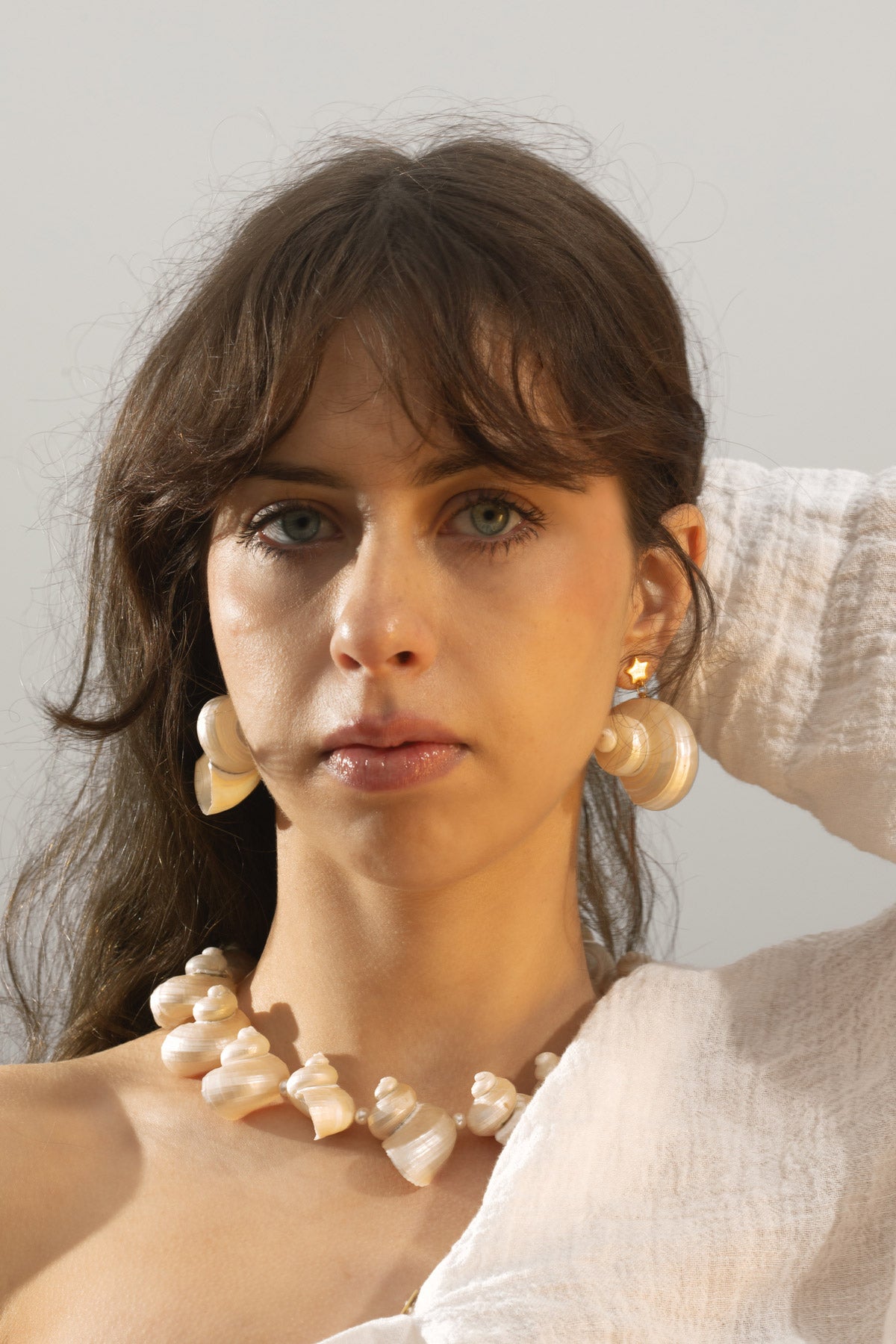 lose-up of a model, holding her hair back as it flows in the wind. Her hair frames the Astr Seashell Dangle Earrings, creating a modern but boho look. She's wearing the Mer Necklace, an artisanal, nature-inspired bold statement piece handcrafted with turbo seashells and freshwater pearls.