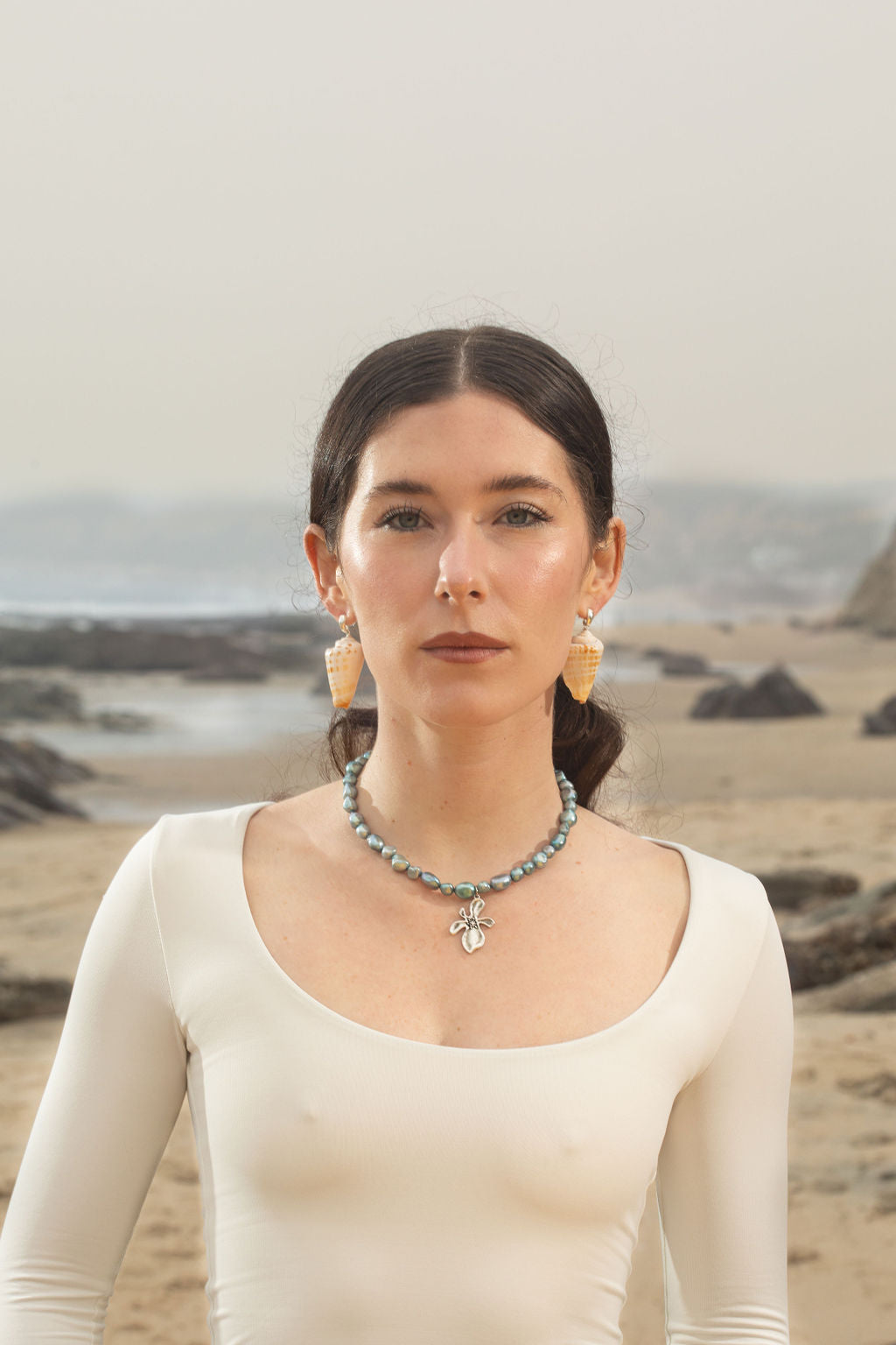 Model wearing the Cleo Necklace, a blue baroque pearl pendant with a silver hibiscus flower, paired with Bea Earrings—sterling silver dome huggies with cone-shaped seashell drops. Her hair is pulled back as ocean waves crash behind her, highlighting the coastal-inspired jewelry.