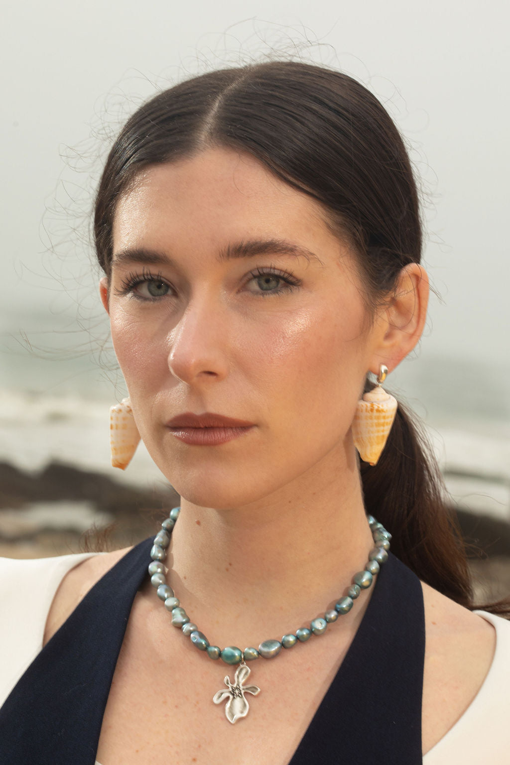 Close-up of the model wearing the Cleo Necklace with chunky blue baroque pearls and a sterling silver hibiscus pendant, paired with Bea Earrings featuring cone-shaped seashell drops. The rocky terrain backdrop emphasizes the ocean-inspired jewelry’s natural textures.