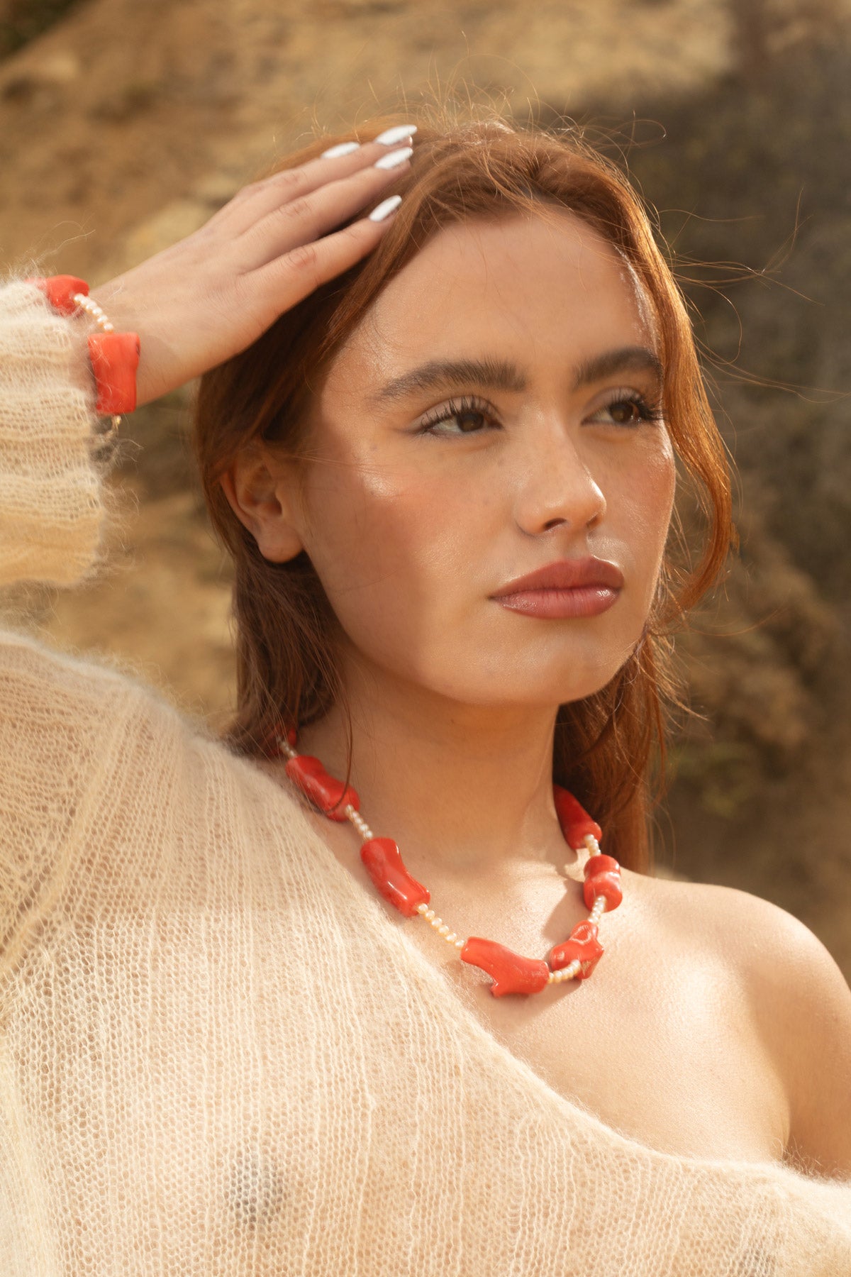 Model wearing the Coral Necklace and its matching Coral Mini Bracelet, both crafted with chunky coral beads and freshwater pearls. Set against a dramatic rocky terrain backdrop, showcasing ocean-inspired jewelry.