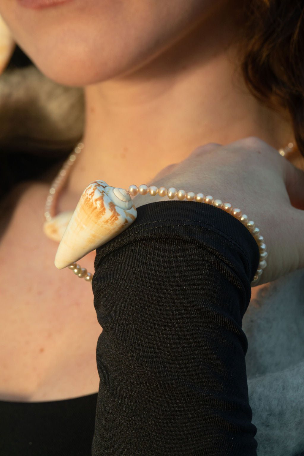 Close-up of a model's arm bent backwards, showcasing the Isla Mini Bracelet. The freshwater pearls and chunky conch shell pendant stand out beautifully against the warm golden hour lighting, highlighting the ocean-inspired design.
