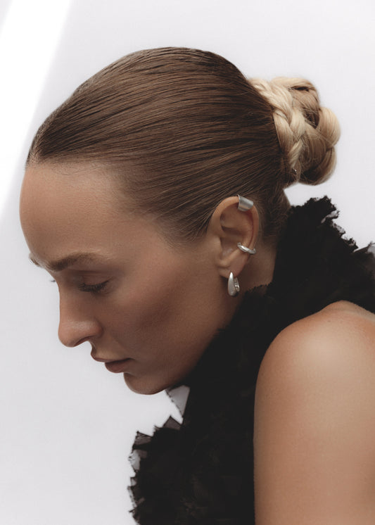 Side profile of a model wearing the Keeper Earrings — elongated sterling silver hoops with a fluid, sculptural shape, styled alongside other silver ear cuffs