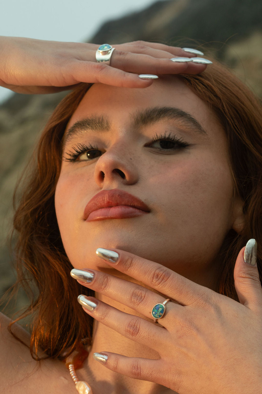 The model, now looking down toward the camera, highlights both the Opal Mosaic I and II Rings, each featuring opal mosaic cabochons set in sterling silver. Her left hand rests gently by her face, showcasing the ocean-inspired Opal Mosaic I Ring, while the Opal Mosaic II Ring adds depth with its chunky, concave design. The Pia Necklace, a beaded freshwater pearl and Condor agate piece, completes the look with a touch of natural beauty. Each handmade piece reflects unique artisanal craftsmanship, perfect for 