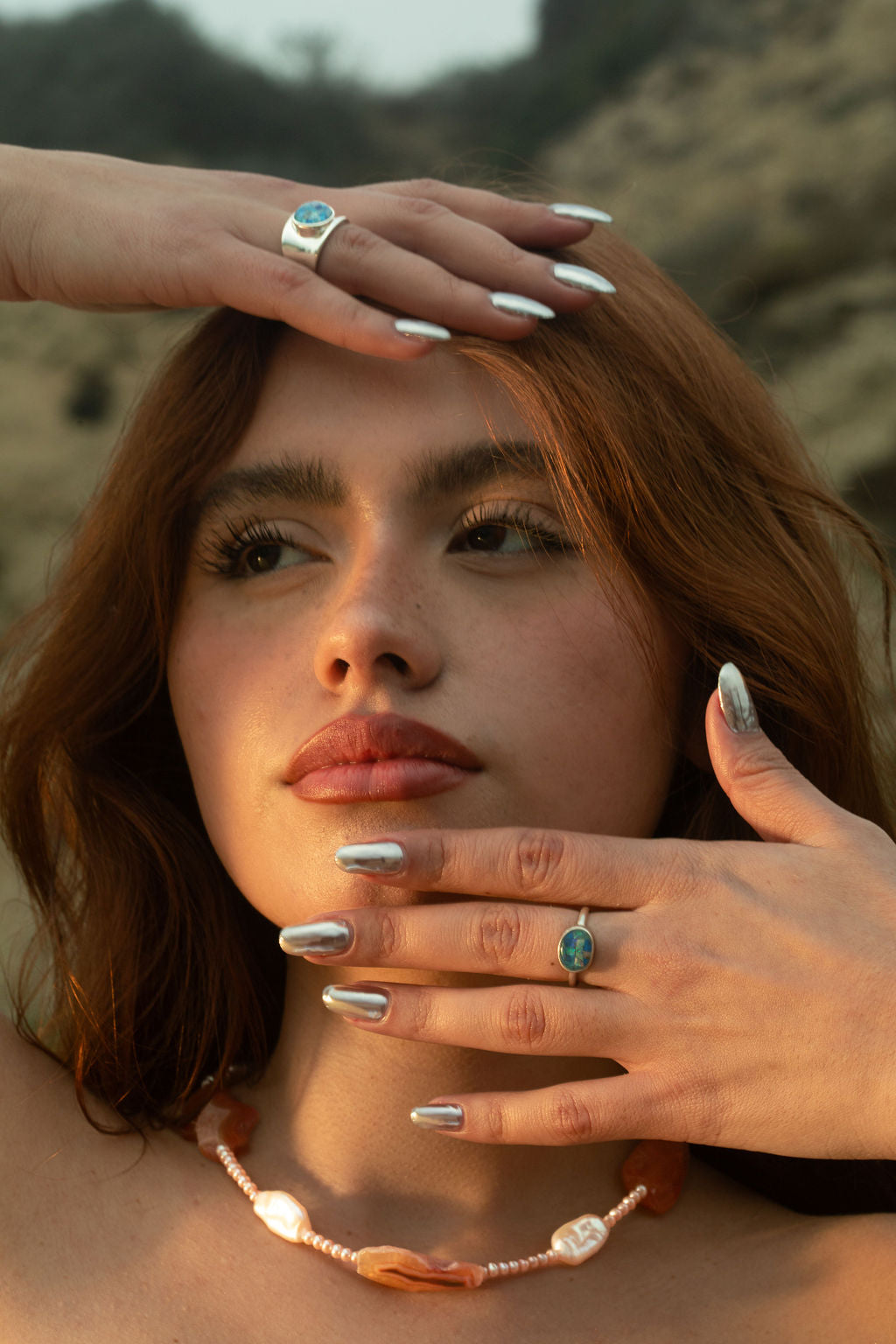 A model's left hand delicately positioned in front of her face, showcasing the Opal Mosaic I Ring, paired with the Opal Mosaic II Ring on her other hand. The opal gemstones stand out against a rocky coastline backdrop. The intricate designs are complemented by the Pia Necklace, a beaded freshwater pearl and Condor agate piece.