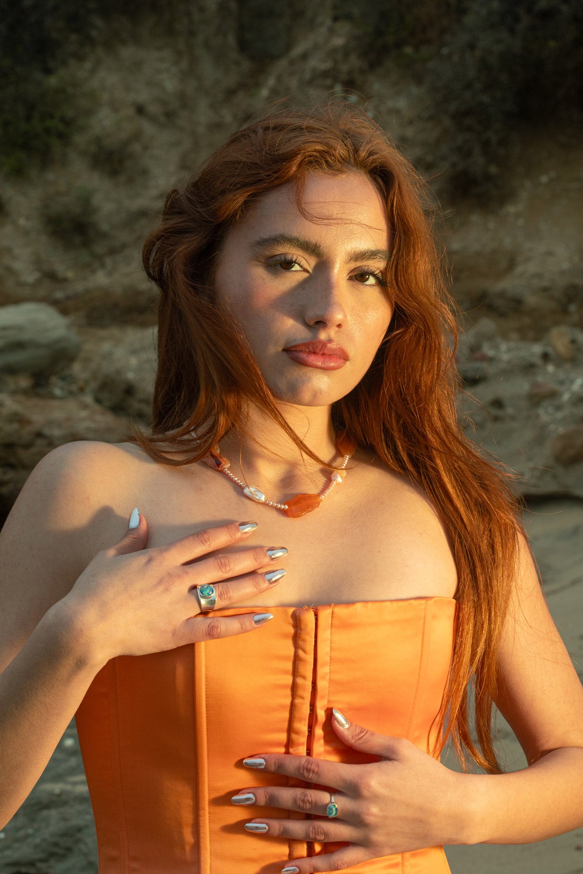 Evoking a modern but soft look, the model stands against a rocky beach hilltop, showcasing the Pia Necklace with Condor agate, freshwater keshi, and baroque pearls. She's also wearing the Opal Mosaic II Ring, a chunky sterling silver concave ring with an opal mosaic cabochon, and the Opal Mosaic I Ring, an oval sterling silver ring with an opal mosaic cabochon. 