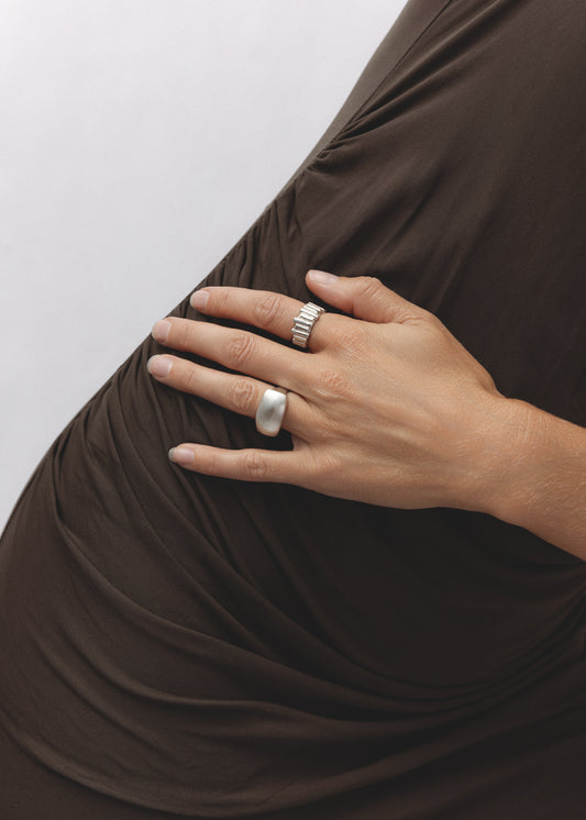 A close-up of a hand resting against ruched brown fabric, wearing the Relic Ring — a textured, organic band with ridged details — stacked alongside a smooth silver dome ring.