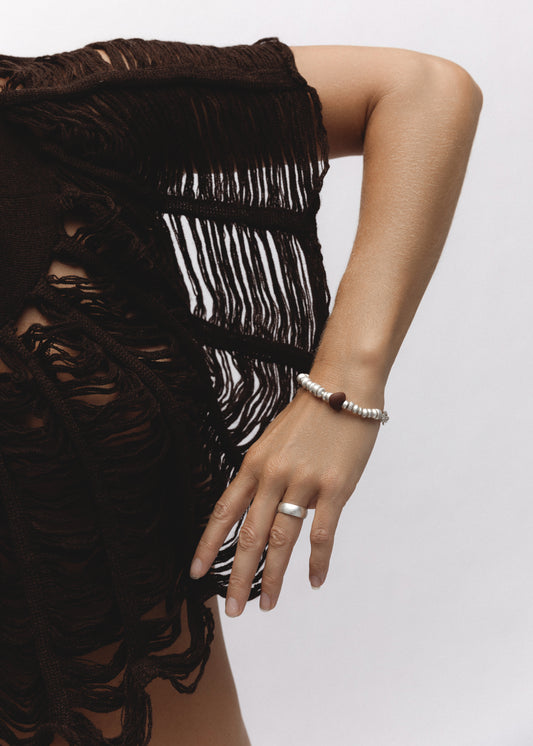 Model wearing the Remnant Bracelet, a strand of irregular sterling silver beads with a single red stone accent, styled on the wrist. The arm rests against the body, dressed in a dark fringed garment, with a silver band ring on the hand.