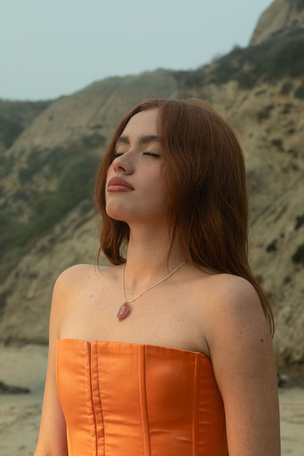 Model wearing the Sol Necklace. The sterling silver chain and Condor Agate pendant illuminate against the rocky beach hillside backdrop.
