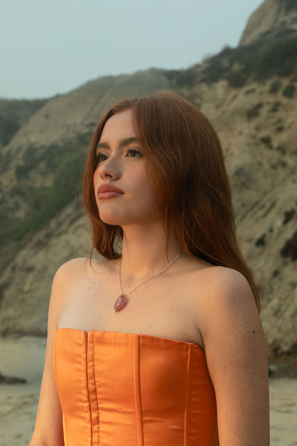 Model wearing the Sol Necklace. The sterling silver chain and Condor Agate pendant illuminate against the rocky beach hillside backdrop. 
