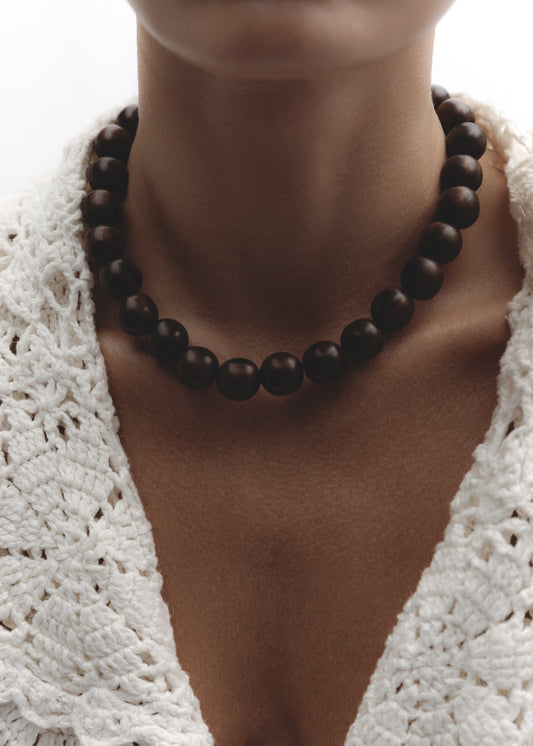 Model wearing the Totem Necklace — bold strand of kamagong wood beads in dark brown, styled against a white crochet top.